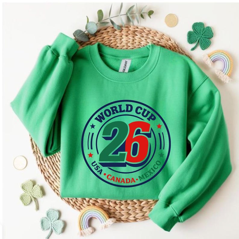 May include: A green sweatshirt with the text "WORLD CUP 26" in a circular design. The design includes the flags of USA, Canada, and Mexico. The sweatshirt has long sleeves and a crew neck.