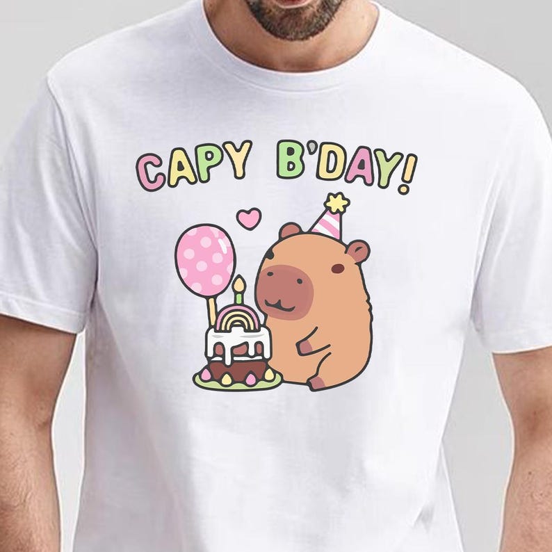 May include: White t-shirt featuring a cartoon capybara with a party hat, a pink balloon, and a birthday cake. The text "CAPY B'DAY!" is written above the capybara in colourful letters. A heart is also included in the design.