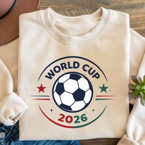 May include: Cream-colored sweatshirt with a circular graphic featuring a soccer ball and the text "WORLD CUP 2026". The design includes red and green accents and star shapes, perfect for soccer fans.