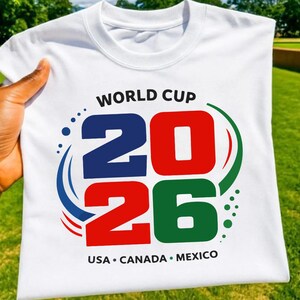 May include: White t-shirt with the text "WORLD CUP 2026" in blue, red, and green. The shirt also features the text "USA &bull; CANADA &bull; MEXICO" in black. The shirt is held up against a green background.