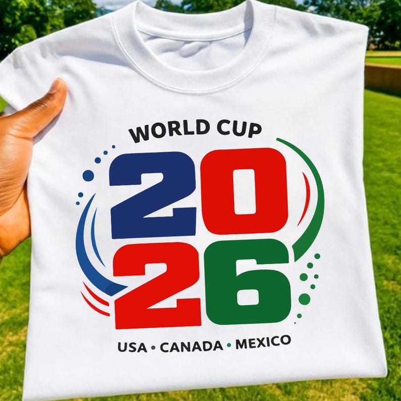 May include: White t-shirt with the text "WORLD CUP 2026" in blue, red, and green. The shirt also features the text "USA &bull; CANADA &bull; MEXICO" in black. The shirt is held up against a green background.