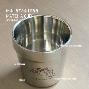 Könnte beinhalten: Ein Mini-Matcha-Becher aus Edelstahl mit 150 ml Fassungsvermögen und doppelwandiger Konstruktion. Die Außenseite zeigt eine Gravur von zwei kleinen Tieren, die durch ein Band verbunden sind, sowie den Text MINI STAINLESS MATCHA CUP ARTE LOUNGE.