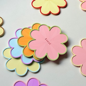 May include: A collection of flower-shaped coasters in various pastel colours including pink, yellow, orange, blue, and purple. Each coaster features a scalloped edge and a contrasting border colour. Some coasters are embossed with names like Leanne and Lowai.