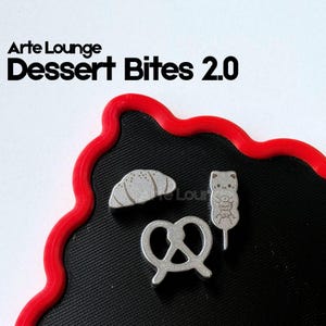 May include: Three silver-toned metal pins featuring a croissant, a pretzel, and a cat-shaped treat on a stick. The text Arte Lounge Dessert Bites 2.0 is printed above the pins. The pins are arranged on a black surface with a red wavy border.