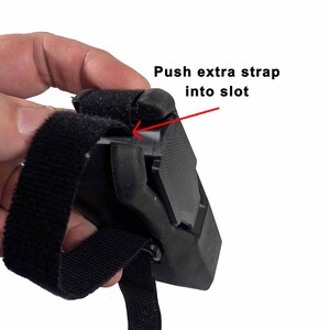 May include: A close-up of a black strap and buckle mechanism. The text "Push extra strap into slot" is visible, with an arrow pointing to the slot. The strap is made of a textured material, and the buckle is a solid, molded piece.