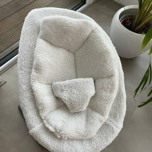 Può includere: Un seggiolino a dondolo per neonati in peluche bianco con una texture morbida e soffice. Il seggiolino ha una seduta profonda e arrotondata e un poggiatesta rialzato.
