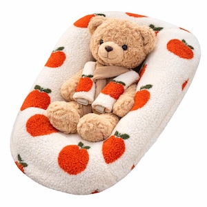 Puede incluir: Un oso de peluche con una bufanda est&aacute; sentado en una cama para mascotas blanca de forma ovalada. La cama tiene un patr&oacute;n de manzanas naranjas. El oso es de color marr&oacute;n claro con una bufanda beige y calentadores de brazos a juego.