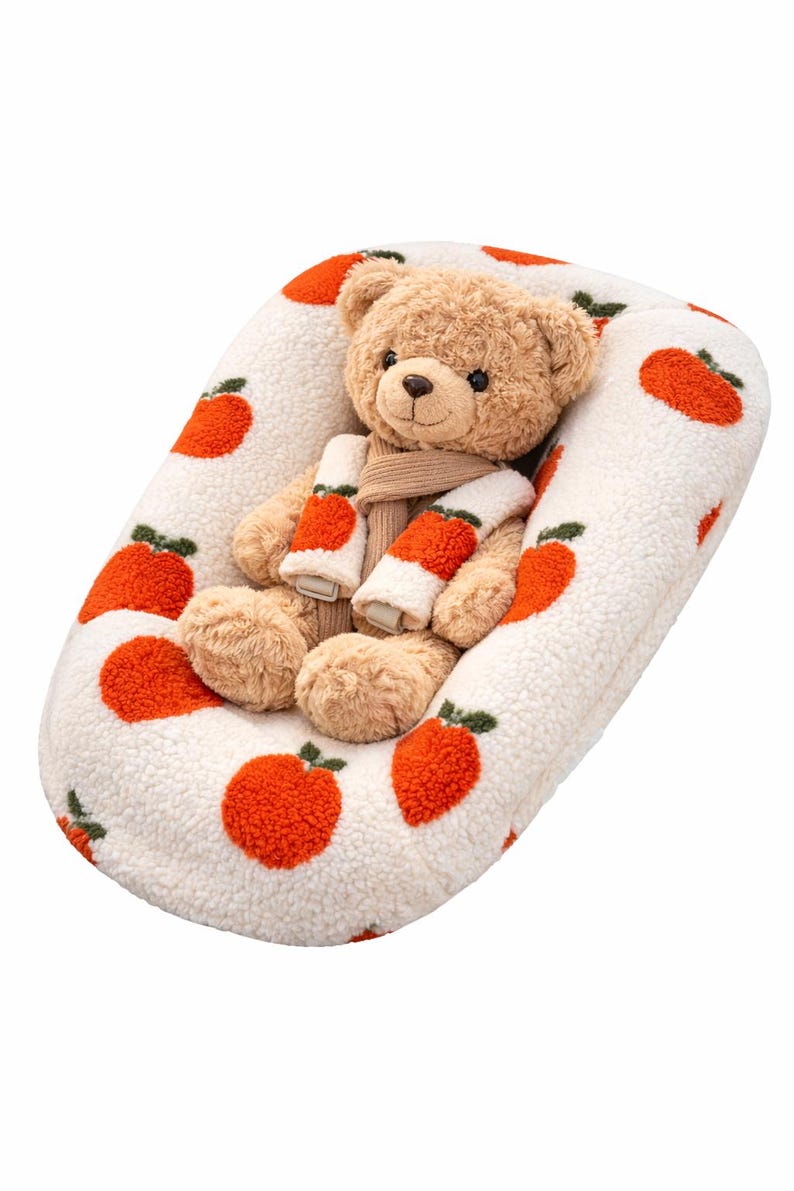 Puede incluir: Un oso de peluche con una bufanda est&aacute; sentado en una cama para mascotas blanca de forma ovalada. La cama tiene un patr&oacute;n de manzanas naranjas. El oso es de color marr&oacute;n claro con una bufanda beige y calentadores de brazos a juego.