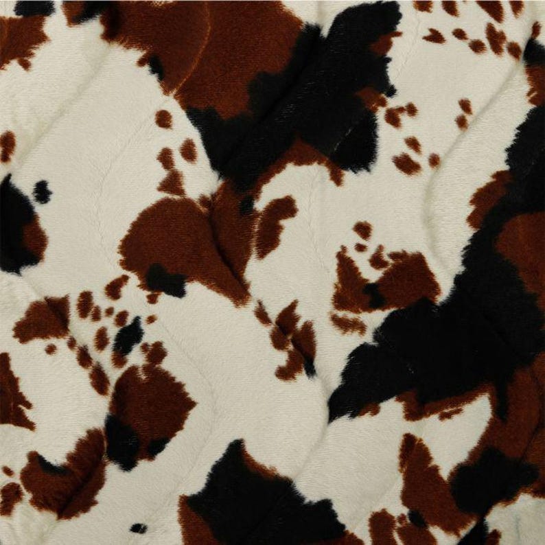 May include: A soft, plush fabric featuring a repeating cow print pattern in shades of cream, dark brown, and black. The material has a visible quilted texture with diagonal stitching lines running across the surface of the fuzzy textile.