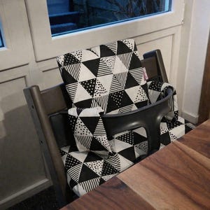 May include: A wooden high chair with a black seat and a patterned cushion. The cushion features a black and white geometric triangle design. The high chair is positioned near a wooden table and a window.