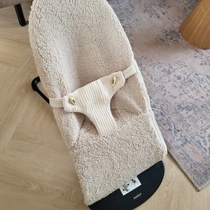 May include: A beige baby bouncer with a plush, textured seat and a waffle-knit safety harness. The bouncer has a black base and a black frame. The bouncer is designed for infants and young children.