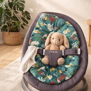 Puede incluir: Una hamaca gris para bebés con un asiento estampado con tucanes, perezosos y otros animales sobre un fondo verde azulado. Un conejito de peluche está sentado en el asiento, sujeto por un arnés gris. Una manta blanca está sobre el lateral. La hamaca está sobre una alfombra clara.