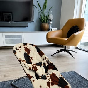 May include: A baby bouncer featuring a quilted fabric cover with a cow print pattern in shades of cream, brown, and black. The bouncer has a curved metal frame and a central fabric restraint strap, designed for infant support and comfort.