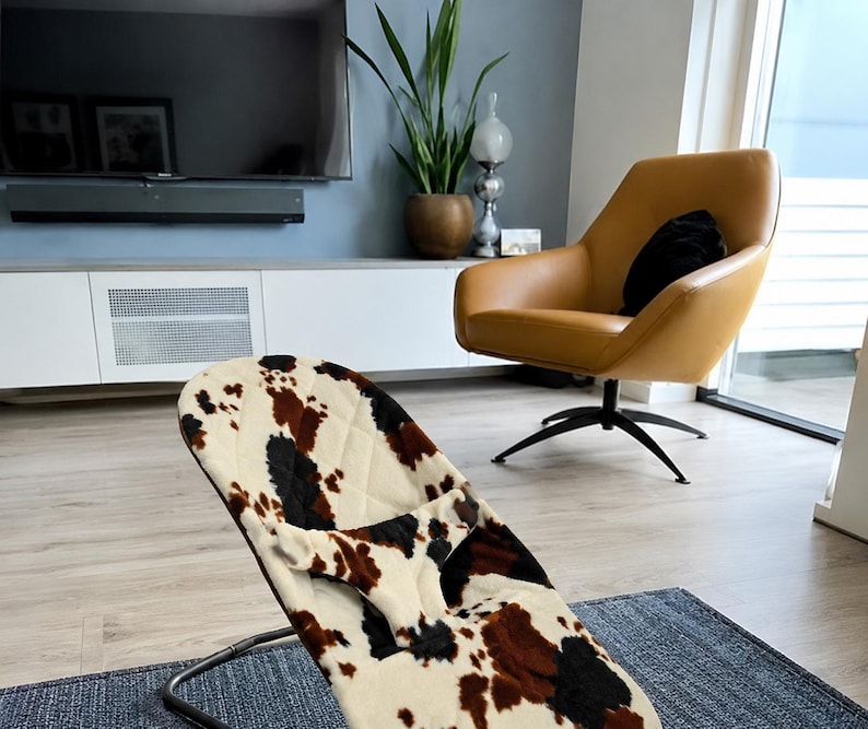 May include: A baby bouncer featuring a quilted fabric cover with a cow print pattern in shades of cream, brown, and black. The bouncer has a curved metal frame and a central fabric restraint strap, designed for infant support and comfort.