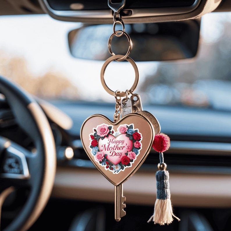 May include: Gold heart-shaped key chain with a pink heart design that says "Happy Mother's Day" surrounded by flowers. The key chain has a tassel and is hanging from a rearview mirror.