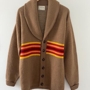 May include: A brown knit cardigan with a shawl collar and a horizontal stripe pattern in yellow, orange, and dark red across the chest. The garment features a button-front closure with dark brown buttons and a Pendleton brand label at the neck.