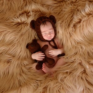 May include: A newborn baby wearing a brown knitted bear hat and holding a brown teddy bear, lying on a brown faux fur blanket.