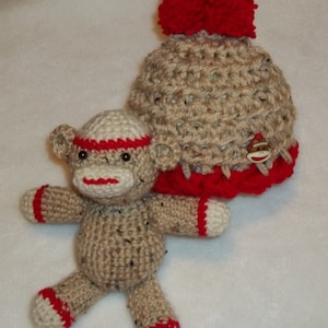 May include: A crocheted monkey toy with red and white accents, wearing a crocheted hat with a red pom-pom and a monkey face button.