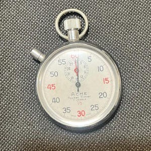 May include: A silver-toned Acme Pocket Timer stopwatch with a white face, black numerals, and red accents. The dial features a central sub-dial, a winding crown with a top ring, and a side pusher. The text reads Acme Pocket Timer 7 Jewels 1/5.