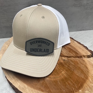 May include: A beige and white trucker hat with a black patch that reads "Overworked and Underlaid".