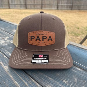 Peut inclure: Un chapeau de camionneur brun et beige avec un patch en cuir brun qui dit "Peyton's Papa Est. 2024". Le chapeau a également une étiquette rouge et blanche qui dit "Richardson 112 Original Trucker Adjustable".