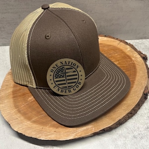 May include: A brown and tan trucker hat with a brown patch that says "One Nation Under God" with a cross and an American flag design.