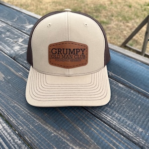May include: A brown and beige trucker hat with a brown leather patch that says "Grumpy Old Man Club" and "Founding Member".