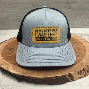 May include: A gray and black trucker hat with a brown leather patch that reads "In my defense I was left unsupervised".