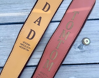 May include: Two leather camera straps with gold-tone metal rivets. The tan strap is engraved with DAD and the names EVAN, WESLEY, and ROWYN. The reddish-brown strap is engraved with TOMTOM and the names RHETT, MILLER, and the text EST. 2018.
