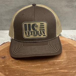 May include: A brown and tan trucker hat with a brown brim and a mesh back. The hat has a black and white American flag design with the text "US Veteran" on it.