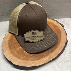 Peut inclure: Casquette de camionneur marron et beige avec un patch marron qui indique "Blue Collar Dollar I wasn't born with it. I worked for it."