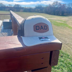 May include: A beige and black trucker hat with a brown leather patch that says "DAD" on it. The hat is sitting on a wooden railing.