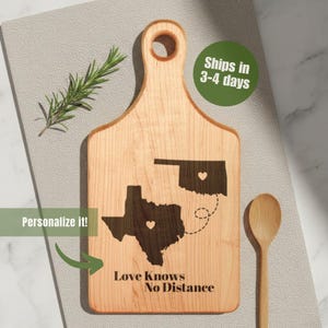 May include: A light-colored wooden cutting board with a handle, featuring engraved outlines of Texas and Oklahoma, connected by a dotted line. The text "Love Knows No Distance" is below the states. A wooden spoon and sprig of rosemary are nearby.