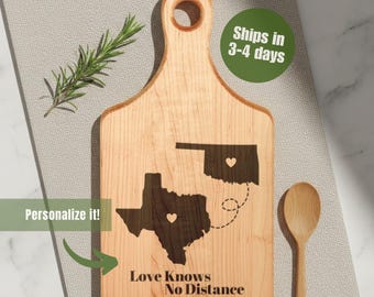 Könnte beinhalten: Ein helles Holzschneidebrett mit Griff, das gravierte Umrisse von Texas und Oklahoma zeigt, die durch eine gestrichelte Linie verbunden sind. Der Text "Love Knows No Distance" steht darunter. Ein Holzlöffel und ein Rosmarinzweig liegen daneben.