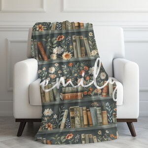 May include: A soft, plush throw blanket featuring a vintage-inspired design of a bookshelf filled with books and flowers. The bookshelf is painted in a dark green color, and the books are a mix of brown, red, and yellow. The flowers are a mix of white, pink, and yellow.