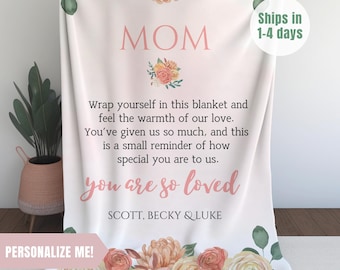 May include: A white throw blanket with a floral design and the text "MOM" in pink. The blanket has a message that reads "Wrap yourself in this blanket and feel the warmth of our love. You've given us so much, and this is a small reminder of how special you are to us. You are so loved." The names "SCOTT, BECKY & LUKE" are printed below the message.