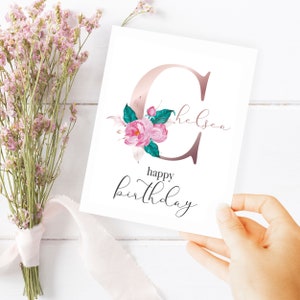 May include: A white card with a pink and green floral design featuring the letter "C" with the name "Chelsea" written in cursive. The text "happy birthday" is written in cursive below the floral design.
