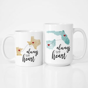 May include: Two white coffee mugs with a long distance relationship theme. One mug features a map of Minnesota, Texas, and Florida with red hearts and the text "always in my heart". The other mug features a map of Ohio and Florida with red hearts and the text "always in my heart".