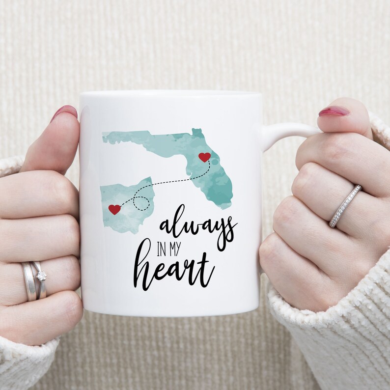 May include: White ceramic mug with a teal watercolor outline of Ohio and Florida with red hearts on each state. A dotted line connects the two states with the text "always in my heart" written in black cursive.