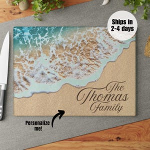 K&ouml;nnte beinhalten: Ein personalisiertes Schneidebrett mit einer Strandszene mit blauem Wasser, wei&szlig;em Schaum und braunem Sand. Der Text "The Thomas Family" ist in Braun auf das Brett gedruckt.