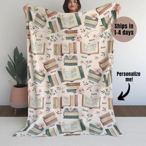 May include: A white fleece blanket with a repeating pattern of open books, stacks of books, and pink flowers. The name "Sara" is repeated throughout the pattern.