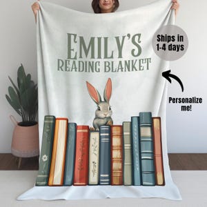 May include: Personalized reading blanket with a white background, the text "Emily's Reading Blanket" in green, and a gray rabbit sitting on a stack of colorful books.
