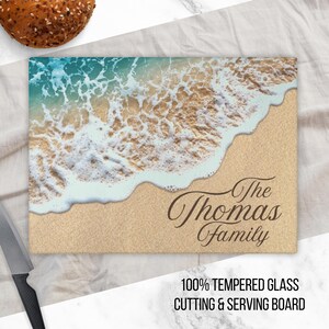 K&ouml;nnte beinhalten: Ein Schneidebrett aus Glas mit einer Strandszene mit blauem Wasser, wei&szlig;en Wellen und braunem Sand. Der Text "The Thomas Family" ist in Braun auf das Brett gedruckt.