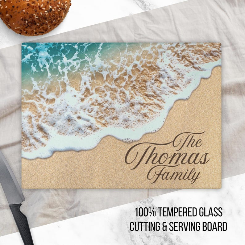 K&ouml;nnte beinhalten: Ein Schneidebrett aus Glas mit einer Strandszene mit blauem Wasser, wei&szlig;en Wellen und braunem Sand. Der Text "The Thomas Family" ist in Braun auf das Brett gedruckt.