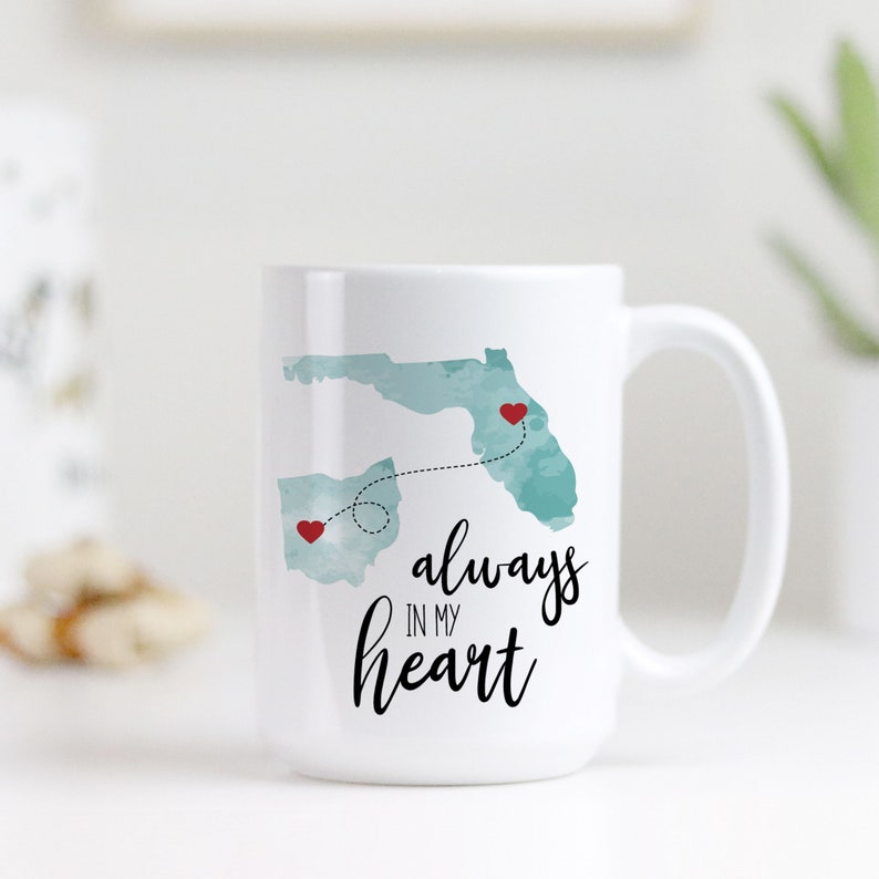 May include: White ceramic mug with a teal watercolor outline of Ohio and Florida with red hearts and a dotted line connecting them. The text "always in my heart" is written in black cursive.