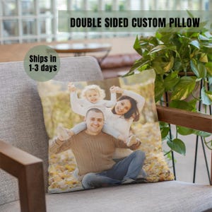 May include: A custom pillow with a photo of a family of three in a park setting. The photo shows a man, a woman, and a young girl. The man is holding the girl on his shoulders, and the woman is standing next to them. The photo is printed on a white pillow with a light brown background.