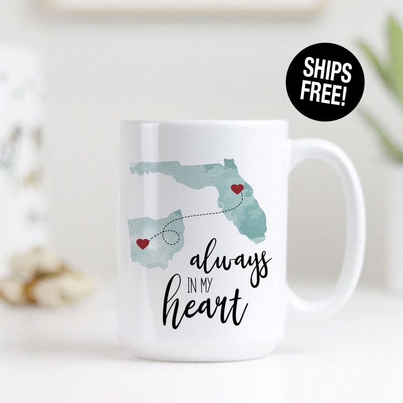 May include: White ceramic coffee mug with a watercolor design of Ohio and Florida with a dotted line connecting the two states. Red hearts are placed on each state. The text "always in my heart" is written in black cursive below the states.
