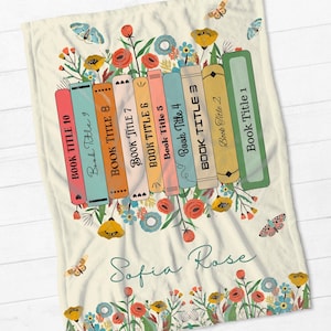 May include: A cream-colored fleece blanket with a stack of colorful books with floral accents and the text "Sofia Rose".