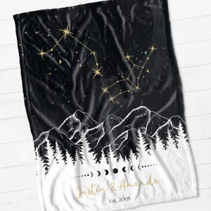 May include: Black and white fleece blanket with a constellation design in gold. The blanket features a mountain range and forest silhouette with the text "Justin & Amanda Est. 2005" in gold.
