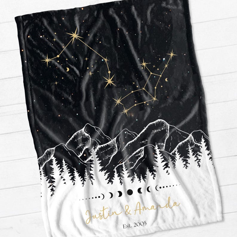 May include: Black and white fleece blanket with a constellation design in gold. The blanket features a mountain range and forest silhouette with the text "Justin & Amanda Est. 2005" in gold.